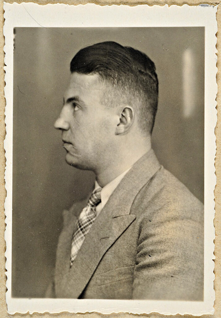 Es ist ein schwarz-weißes Fotoporträt von Herbert Lange. Es hat eine helle Umrandung. Lange ist von der Seite aufgenommen und blickt nach links aus dem Foto heraus. Er hat eine Narbe am Mundwinkel, vermutlich ein sogenannter Schmiss. Er trägt die Haare zurückgekämmt und trägt einen hellen Anzug aus strukturiertem Stoff mit einer karierten Krawatte zum weißen Hemd.