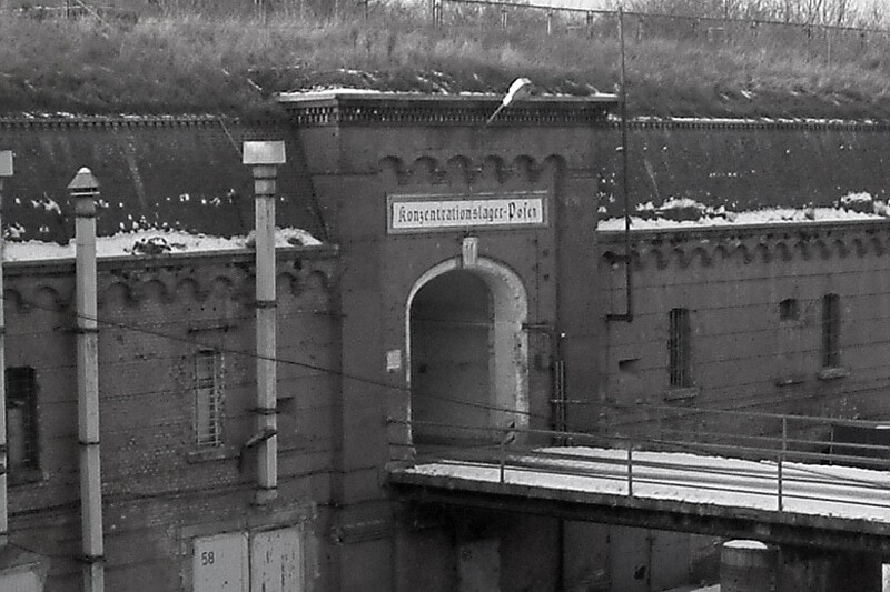 Es ist ein schwarz-weißes Foto eines Eingangs einer Festung. Sie ist mit Ziegelsteinen in einen Verteidigungswalls gebaut. Ein Torbogen ist der Eingang. Über dem Eingang steht in Frakturschrift »Konzentrationslager Posen«. Eine Brücke führt auf das Tor zu, da es sich im oberen Stockwerk befindet. Das Dach des Gebäudes ist mit Gräsern bewachsen.