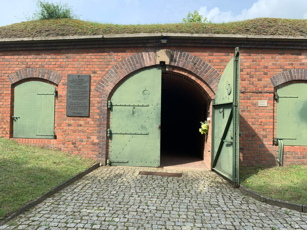 Es ist ein Farbfoto aus dem Inneren der Festung. Abgebildet ist ein Gebäudeabschnitt der aus rotem Backstein geziegelten Festung mit einem hellgrün gestrichenem Stahltor mit Rundbogen. Rechts und links von dem Stahltor sind Fenster, die mit Stahltüren - ebenfalls hellgrün gefasst - verschlossen sind. Der rechte Flügel des Stahltores steht offen, der dahinter liegende Stollen ist dunkel. Links vom Tor ist eine Bronze angebracht. Die Inschrift der Gedenktafel ist in Polnisch, Deutsch und Englisch. Sie lautet: »An diesem Ort verübten die deutschen Besatzer im Oktober 1939 den ersten Massenmord an psychisch Kranken durch Vergasen.« Über der Inschrift ist ein christliches Kreuz. Die Buchstaben sind erhaben.