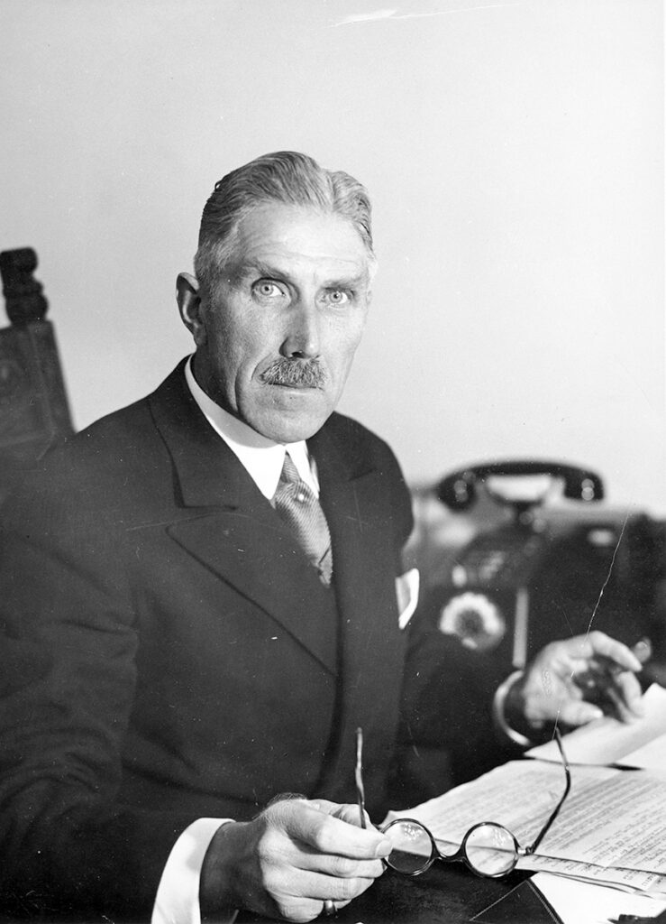 Das ist ein schwarz-weiß Porträtfoto. Franz von Papen blickt mit strengem Blick direkt in die Kamera. Er trägt zurückgekämmte Haare und einen gestutzten Schnurrbart. Außerdem trägt er einen dunklen Anzug mit hellem Hemd und Krawatte.