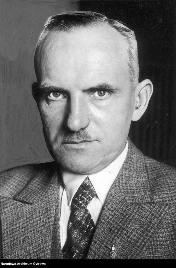 Das ist ein schwarz-weiß Porträtfoto. Arthur Gütt blickt mit geschlossenem Mund und starrem Blick in die Kamera. Er trägt einen kleinkarierten Anzug aus grobem Stoff mit hellem Hemd und strukturierter Krawatte. Sein Haar ist gescheitelt und zurückfrisiert. Das Bild ist mit einem Kohlestift nachbearbeitet worden.