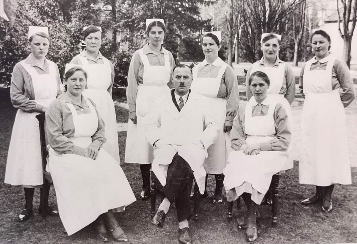 Es ist ein schwarz-weißes Gruppenbild. In der Mitte sitzt Max Bräuner. Er trägt ein Hitler-Bärtchen und die dunklen Haare nach hinten gebürstet. Er trägt einen weißen Arztkittel, darunter sind ein weißes Hemd, eine Krawatte und eine dunkle Weste erkennbar. Er hat die Beine übereinandergeschlagen. Er trägt schwarze Schnürschuhe und weiße Socken. Er hat seine Hände in den Taschen seines Kittels verborgen. Rechts und links neben ihm sitzen zwei uniformierte Pflegerinnen, mit gestreifter Bluse, weißer Schürze und weißer Haube. Sie haben ihre Beine über Kreuz und die Hände gefaltet im Schoß. Hinter ihnen und Max Bräuner stehen sechs weitere Pflegerinnen in gleicher Uniform. Sie blicken ernst in die Kamera, nur eine Pflegerin lächelt verhalten. Die Aufnahme entstand im Freien.