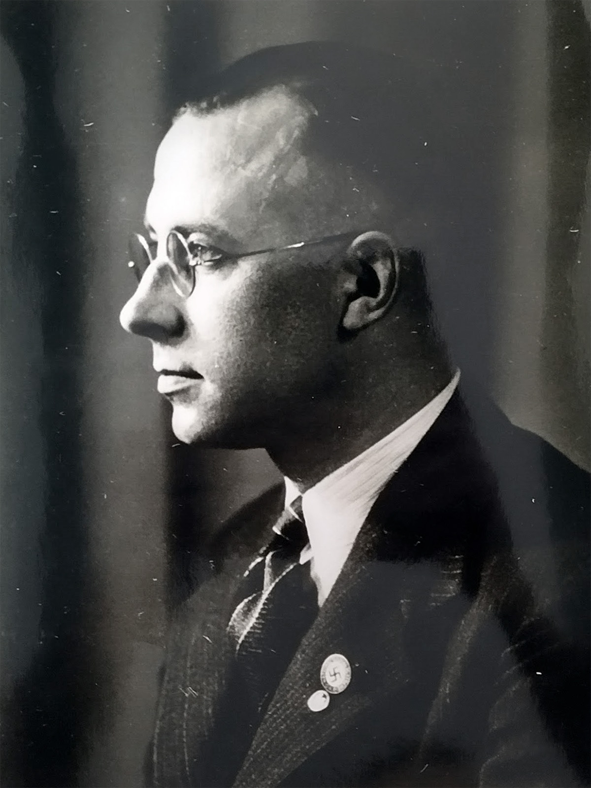 Es ist ein schwarz-weißes Porträt von Willi Baumert. Er sitzt seitlich zur Kamera und blickt links aus dem Bild heraus. Er trägt einen dunklen Anzug mit hellem Hemd und Krawatte. Am Revers trägt er das Pateiabzeichen der NSDAP mit Hakenkreuz. Er hat dunkle, kurze Haare nach hinten gekämmt und trägt eine Nickelbrille.