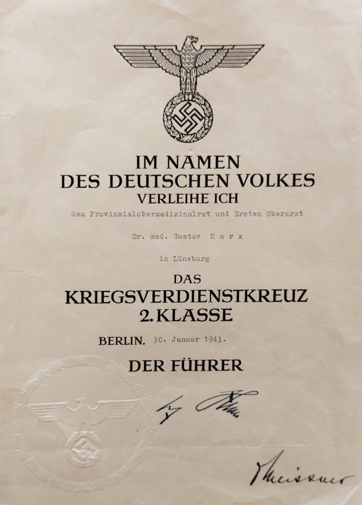 Es ist vergilbtes Papier. Es zeigt den Reichsadler, der einen Kranz aus Eichenblättern in den Klauen hat, in dessen Mitte ein Hakenkreuz ist. Darunter steht in großen Buchstaben: Im Namen des deutschen Volkes verleihe ich. Es folgt in Maschinenschrift geschrieben: dem Provinzialobermedizinalrat Dr. med. Gustav Marx in Lüneburg. Es folgt in großen Buchstaben: Das Kriegsverdienstkreuz 2. Klasse. Berlin. Es folgt in Maschinenschrift: 30. Januar 1943. Es folgt in großen Buchstaben: Der Führer. Darunter befindet sich in Tinte die Unterschrift von Adolf Hitler. Unten rechts am Blattrand steht eine zweite Unterschrift: O. Meissner. Es ist die Unterschrift des Staatssekretärs in der Präsidialkanzlei. Das Papier trägt unten links eine Prägung. Es ist das Emblem mit Reichadler, Eichenkranz und Hakenkreuz.
