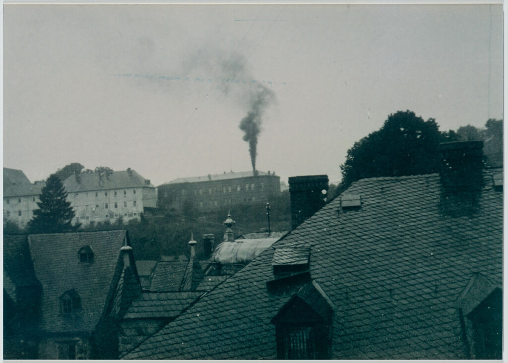 Es ist ein schwarz-weißes Foto. Es ist eine Aufnahme aus einem oberen Stockwerk auf die Heil- und Pflegeanstalt Hadamar in der Ferne. Hinter dem Hauptgebäude der Anstalt steigt dunkler Rauch in den Himmel. Es ist der Rauch des Krematoriums. Im Vordergrund des Bildes sind mit Schiefer gedeckte Dächer von Wohnhäusern zu erkennen. Es scheint geregnet zu haben.
