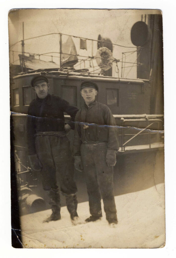 Das Foto ist schwarz-weiß. Im Hintergrund ist ein Schiff zu erkennen. August Golla steht neben einem älteren Mann an der Hafenkante. Er trägt dunkle Arbeitskleidung und feste Handschuhe. Auf dem Kopf hat er eine Mütze. Am Boden und an seinen Schuhen ist etwas Schnee zu sehen.