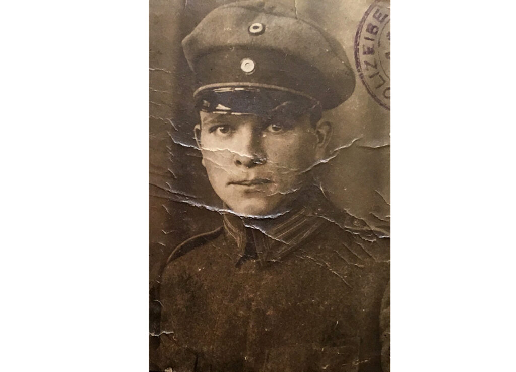 Es ist ein schwarz-weißes Porträtfoto. Es ist sehr klein und stark zerknittert. Ein junger Mann in dunkler Uniform und Schirmmütze ist bis zur Brust zu sehen. Er steht ein wenig seitlich und blickt freundlich direkt in die Kamera. Oben rechts auf dem Foto ist ein Teil eines Stempelabdrucks zu sehen.