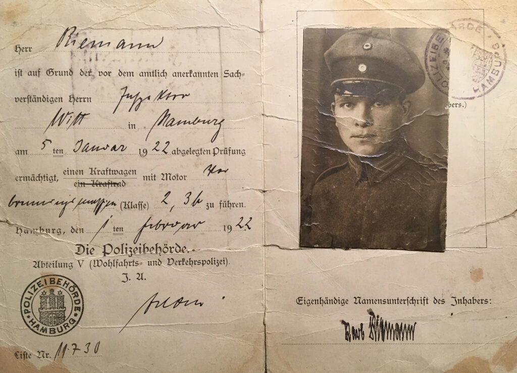 Der Führerschein von Carl Riemann ist aus Papier, mittig gefaltet. Er ist vergilbt und wirkt mitgenommen durch viele Knicke und Risse. Links sind die Angaben im gedruckten Formular handschriftlich ausgefüllt. Leicht schief ist auf die rechte Seite ein Passfoto aufgeklebt. Auf dem Foto trägt Carl Riemann eine hochgeschlossene dunkle Polizeiuniform. Er blickt mit freundlichem Blick in die Kamera.
