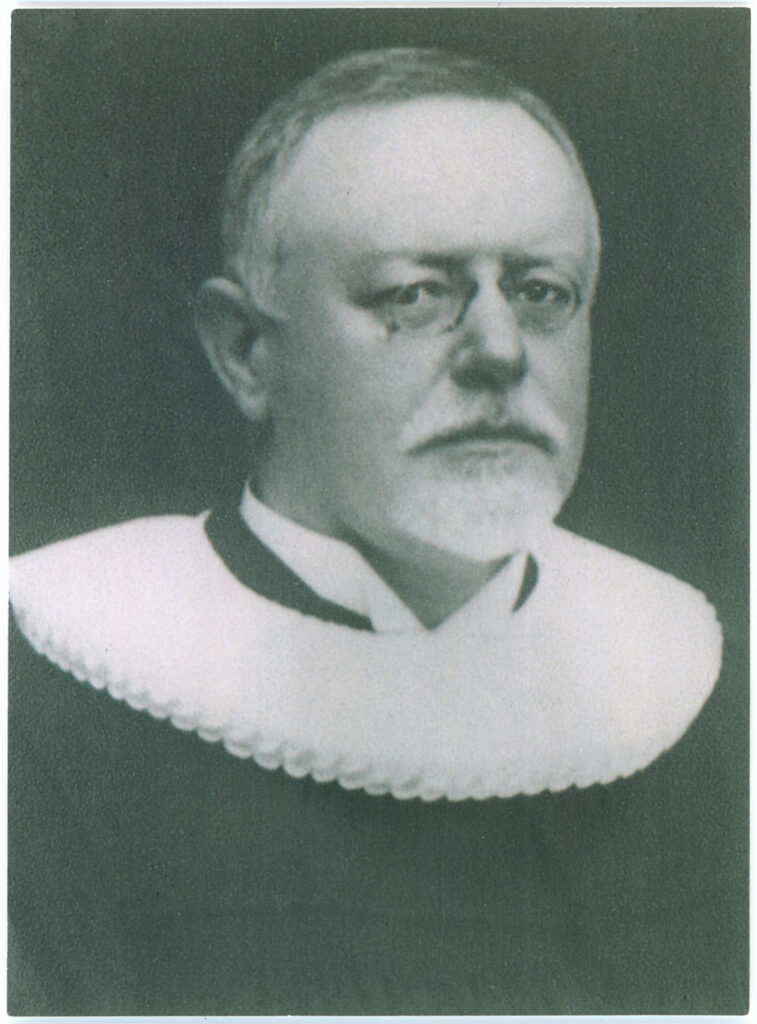 Es ist ein schwarz-weißes Porträt von Heinrich Mund. Er trägt einen Pastorentalar mit weißem, "Hamburger" Kragen. Er trägt eine runde Brille mit Metallgestell. Mit ernstem Blick schaut er direkt in die Kamera. Er hat eine hohe Stirn und trägt einen ergrauten Kinnbart. Auf dem Bild ist er über 60 Jahre alt.