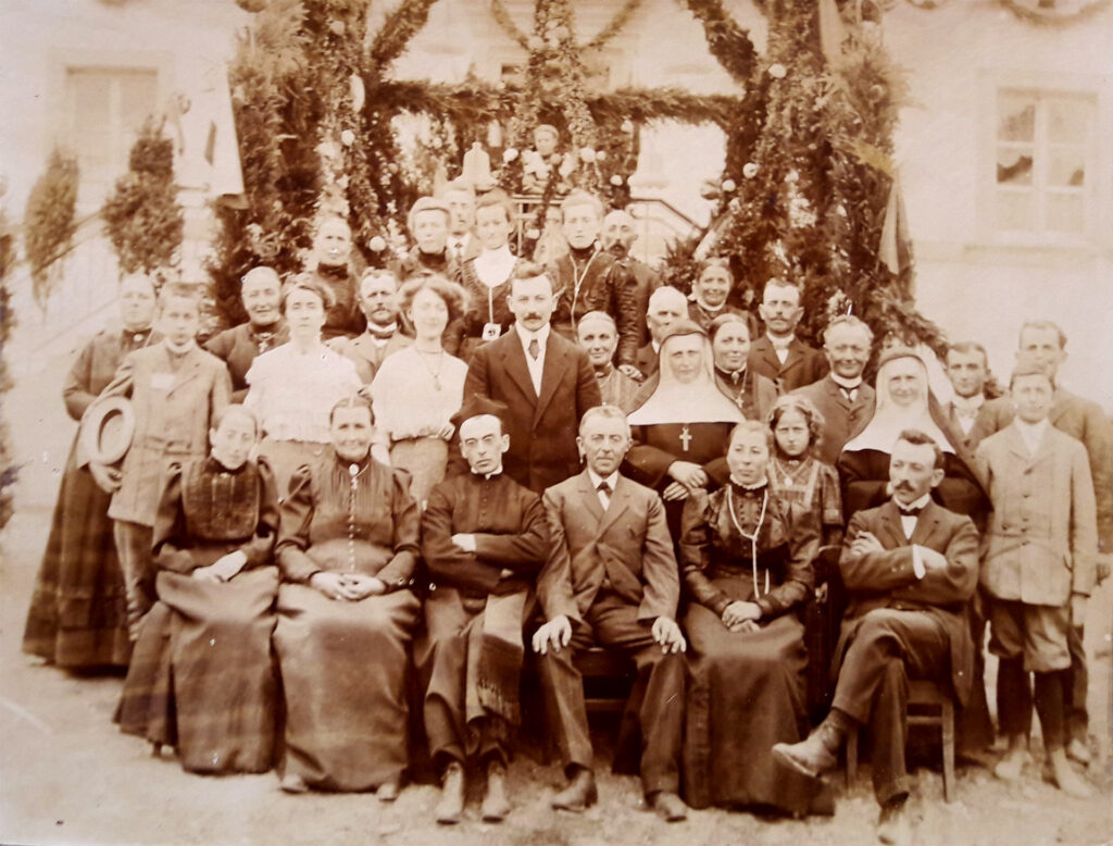 Es ist ein Gruppenfoto und zeigt 31 Personen. Die vorderen Personen sitzen. Die dahinter stehen. Es sind Männer und Frauen, Kinder und alte Menschen. Zwei Nonnen sind darunter. Elsa Spartz steht mittig in der Gruppe und trägt eine helle Bluse sowie einen dunkleren Rock. Heinrich Spartz steht neben ihr und trägt einen dunkeln Anzug mit Weste und dunkler Krawatte. Er trägt einen Schnurrbart und kurze, nach hinten gekämmte Haare.