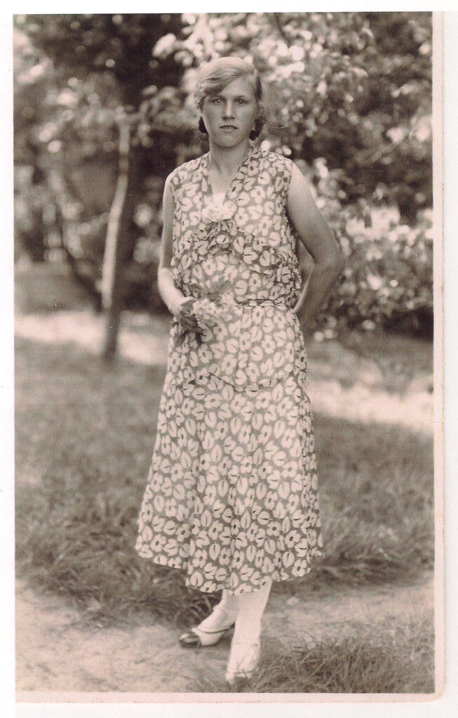 Agnes steht in einem Garten. Sie trägt ein langes, kurzärmeliges Kleid mit großem Blumenmuster. In der rechten Hand hält sie einen kleinen Strauß Blumen. Sie blickt ernst direkt in die Kamera. Ihre Haare sind nach hinten frisiert.