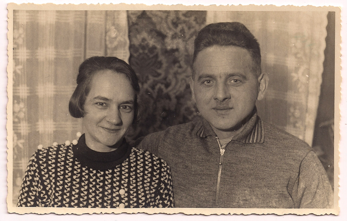 Es ist ein schwarz-weißes Foto. Bertha und Christian Ossmer sind bis zur Brust zur sehen. Christian hat seinen rechten Arm um Bertha gelegt. Bertha trägt einen schwarz-weiß-gemusterten Strickpullover. Ihre Haare sind gescheitelt. Christian trägt einen Pullover mit Reißverschluss und kurzgeschnittene Haare. Beide blicken mit einem leichten Lächeln in die Kamera.