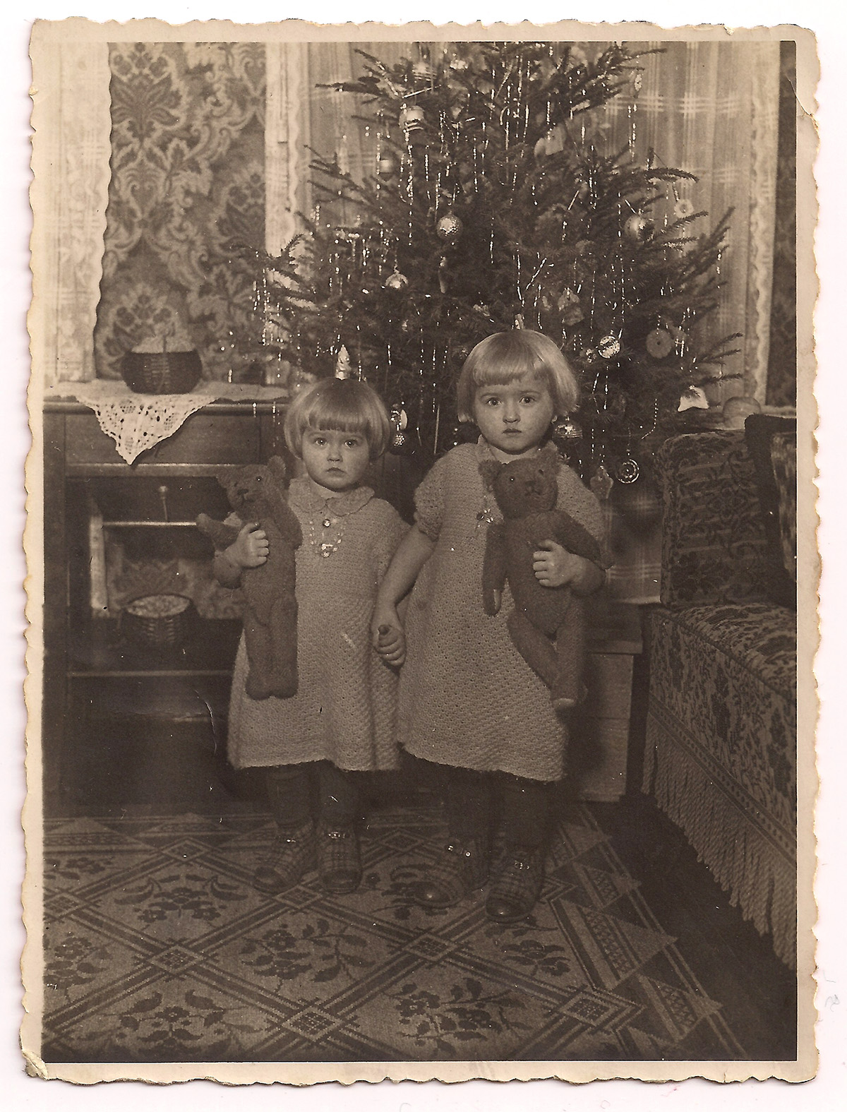 Es ist ein schwarz-weißes Foto. Käthe und Elfriede Ossmer stehen vor einem geschmückten Tannenbaum. Sie halten sich an den Händen. In den anderen Händen tragen beide einen Teddybären. Sie sind identisch gekleidet mit karierten Hausschuhen, dunklen Strumpfhosen und Strickkleidern. Ihre hellen Haare sind zu einem Pagenschnitt frisiert. Die beiden sehen einander sehr ähnlich.