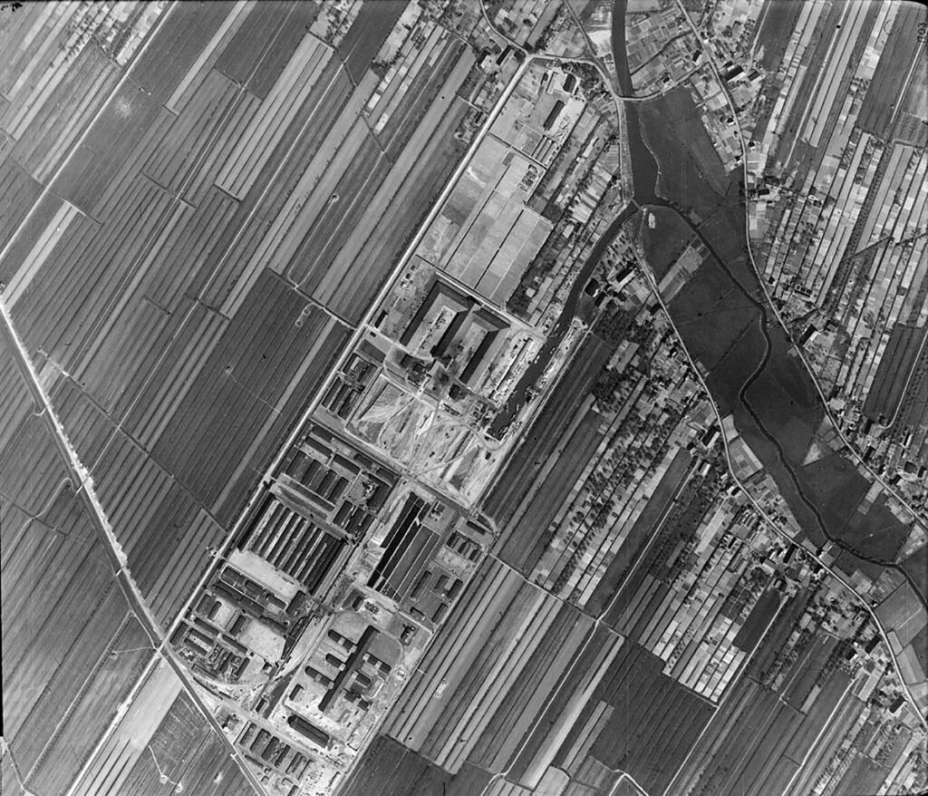 Die Luftaufnahme zeigt das Gelände von Neuengamme sowie die Umgebung. Deutlich zeichnet sich die Struktur der Gebäude des Lagers vor den landwirtschaftlichen Betrieben entlang einer Straße mit dahinterliegenden landwirtschaftlichen Flächen ab.