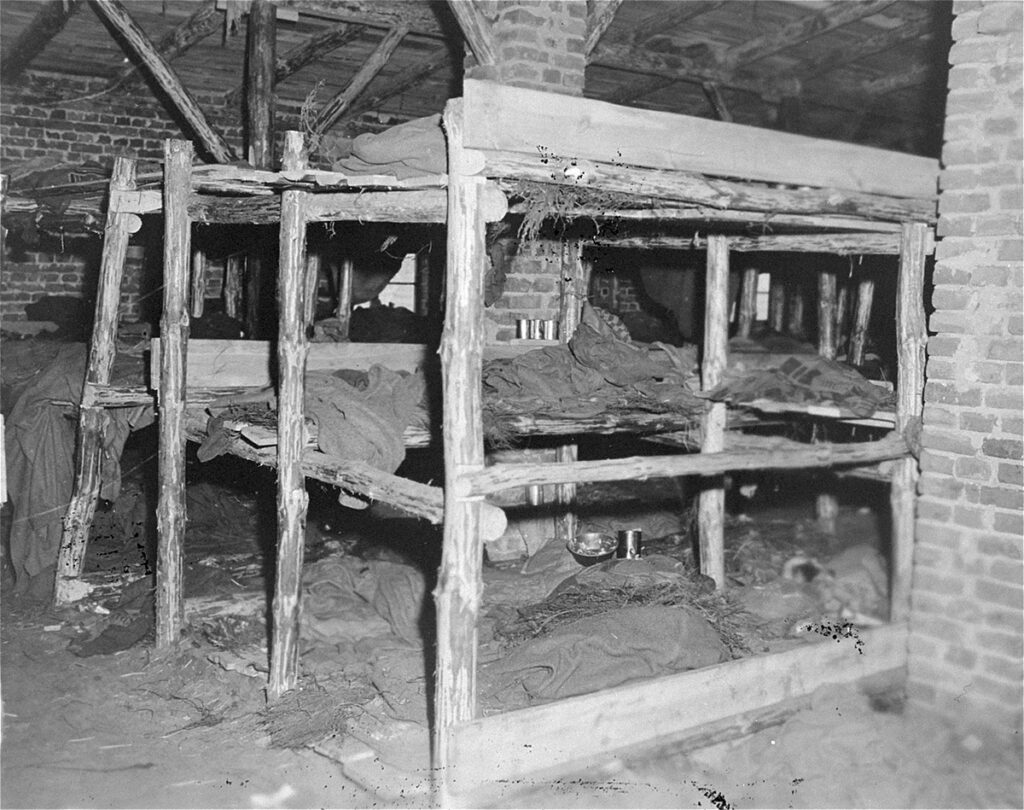 Das schwarz-weiße Foto zeigt einen Blick in eine Schlafstätte. Das dreistöckige Bett steht in einem stallartigen Gebäude. Das Bett hat drei dünne Ebenen, teilweise mit Stroh und groben Stoffen belegt. Der Bettrahmen ist aus grob behauenen Holzstämmen gebaut.