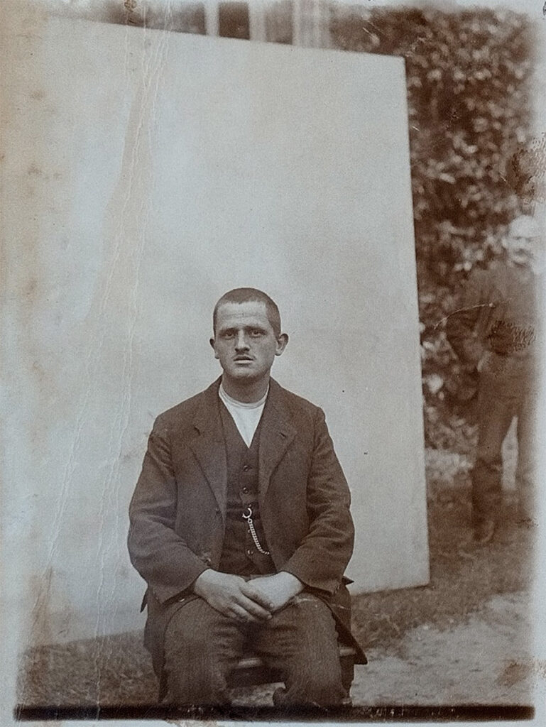 Das Bild zeigt Karl Reich sitzend auf einem Stuhl. Er sitzt vor einer großen hellen Wand. Er trägt einen dunklen Anzug mit Weste und hellem Hemd. Die Kette einer Taschenuhr ist sichtbar. Er hat kurze Haare und blickt mit leicht geöffnetem Mund Richtung Kamera. Seine Hände sind im Schoß gefaltet.