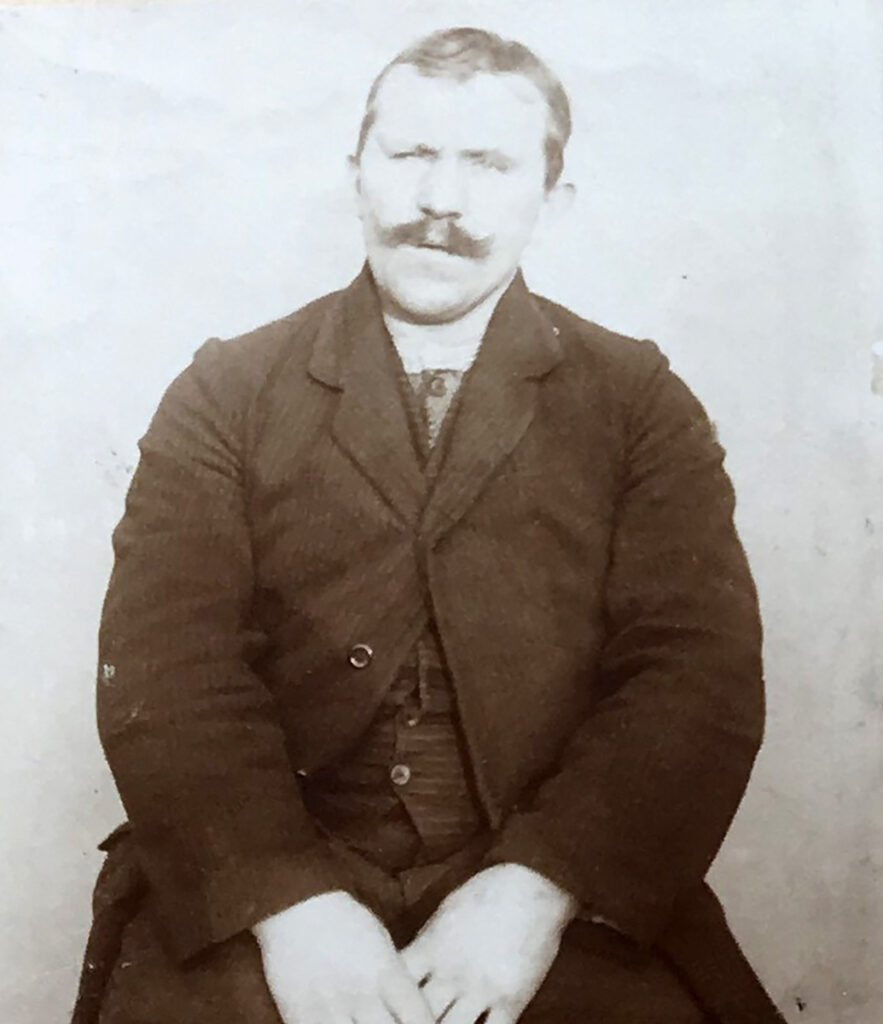 Das Bild zeigt Heinrich Brandt sitzend auf einem Stuhl. Er ist von den Knien aufwärts zu sehen. Er sitzt vor einer großen hellen Wand. Er trägt einen dunklen Anzug mit Weste und hellem Hemd. Er hat kurze, etwas lichte Haare und einen Schnurrbart. Er blickt mit leicht geöffnetem Mund Richtung Kamera. Seine Hände liegen im Schoß.