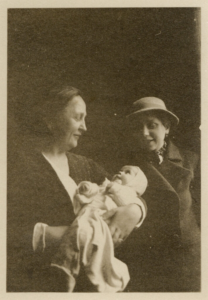 Das Foto ist sepiafarben und insgesamt recht dunkel. Dorothea Kaliwe hält ein Baby in den Armen. Das Baby trägt einen hellen Strampler und blickt aufmerksam zu Dorothea auf. Dorothea trägt dunkle Kleidung und blickt lächelnd auf das Baby herab. Rechts neben den beiden steht Dorotheas Tochter Ursula. Sie trägt einen Damenhut und einen dunklen Mantel mit einem Halstuch. Sie blickt lächelnd auf das Baby herab.