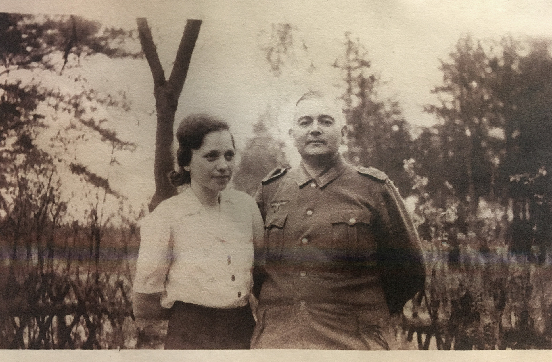 Es ist ein Schwarz-Weiß-Foto. Ursula und Theo Zobel stehen im Freien vor einem hüfthohen Holzzaun. Dahinter befinden sich Bäume und vielleicht ein Gewässer. Beide sind etwa von der Hüfte aufwärts zu sehen. Ursula trägt eine helle, kurzärmelige Bluse und einen dunklen Rock. Ihre Haare sind dunkel und eher kurzgeschnitten. Ihre Arme sind hinter dem Rücken verschränkt. Sie steht ein wenig zur Seite gedreht ihrem Ehemann zugewandt. Sie blickt mit einem leichten Lächeln nach rechts aus dem Bild heraus. Theo steht ein wenig nach links gedreht Ursula zugewandt. Er trägt eine dunkle Uniform und hat die Arme ebenfalls hinter dem Rücken verschränkt. Sein Haar ist kurzgeschnitten und ordentlich gekämmt. Er sieht mit freundlichem Blick direkt in die Kamera.
