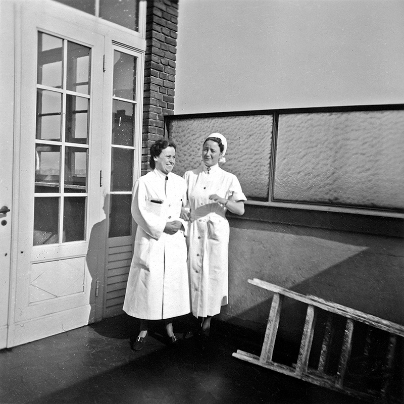 Es ist ein schwarz-weißes Foto. Helene Sonnemann und Harriet Stoltenberg stehen vor einer weißen Tür mit eingesetzten Glasscheiben. Helene Sonnenberg trägt einen weißen Arztkittel und lacht. Ihre dunklen Haare sind wellig. Sie trägt eine Art geöffneten Umschlag unter dem Arm. Sie blickt nach rechts. Harriet Stoltenberg trägt eine Schwestern-Uniform mit einer weißen Haube. Sie hat sich bei Helene Sonnemann untergehakt und lächelt in ihre Richtung. Sie scheint gerade etwas Unterhaltsames gesagt zu haben.