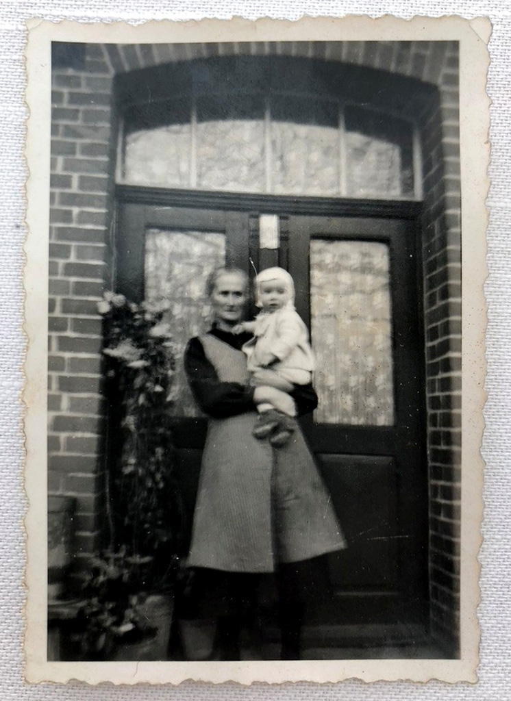 Es ist ein verschwommenes schwarz-weiß Foto. Erich und seine Großmutter stehen vor der Haustür eines Backsteinhauses. Sie trägt einen Kittel über einem dunklen Kleid. Erich ist auf ihrem Arm und trägt helle Kleidung mit einer Babyhaube. Beide blicken in Richtung Kamera.