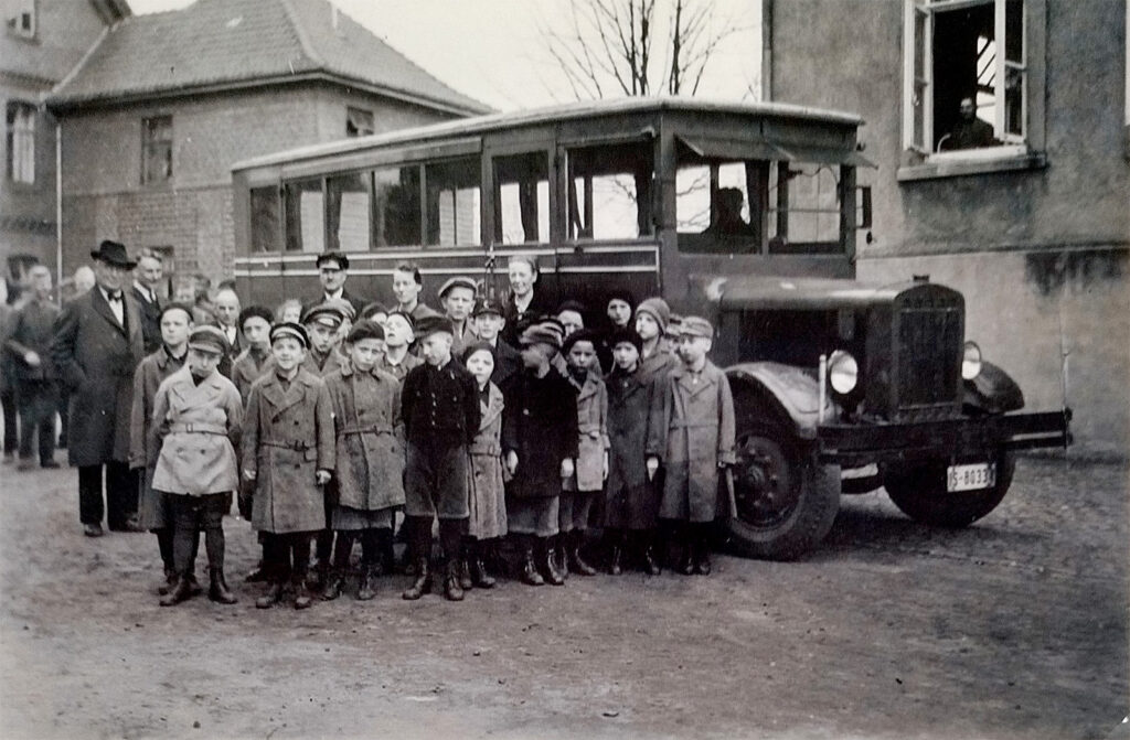 Es ist ein schwarz-weißes Foto. Eine Gruppe von 20 Kindern steht aufgereiht vor einem Omnibus. Die Kinder tragen Mäntel und Mützen. Sie gucken in die Kamera. Hinter ihnen stehen Erwachsene.