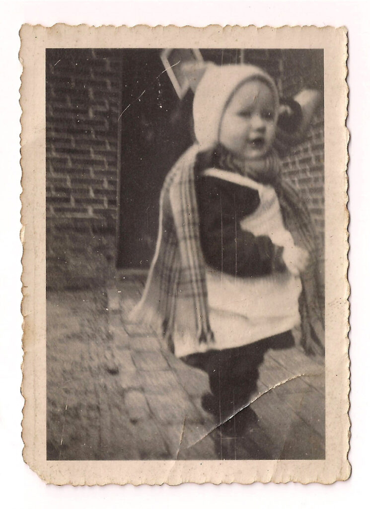 Das ist ein schwarz-weißes Foto. Herta Ley trägt ein Latzkleid und einen karierten Schal. Dazu eine helle Mütze. Sie ist von der Seite aufgenommen und wird an der linken Hand von einer größeren Person gehalten.