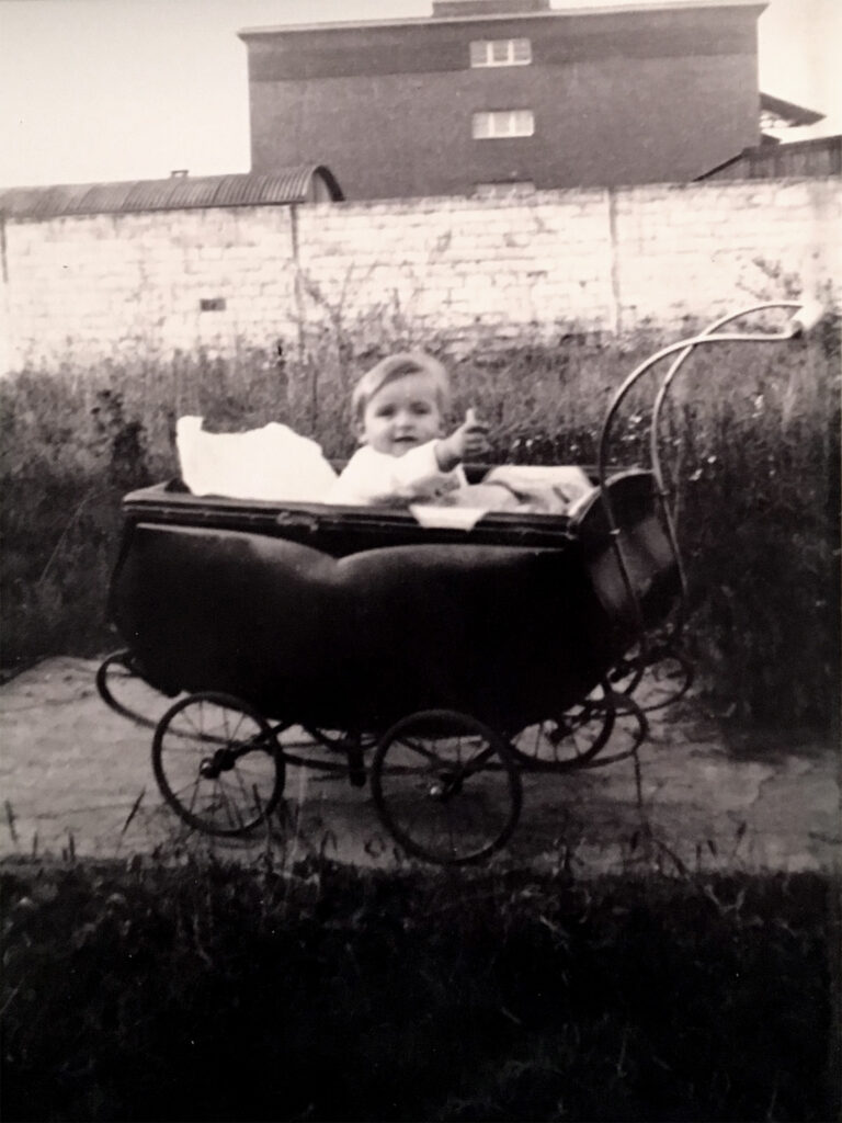 Es ist ein schwarz-weißes Foto. Ingeborg Wahle sitzt in einem ausladenden Kinderwagen mit großen Speichenrädern und geschwungenem Lenker. Sie streckt die rechte Hand aus dem Wagen und blickt Richtung Kamera. Im Hintergrund ist Wiese, eine Mauer und ein dahinterliegendes mehrstöckiges Gebäude sichtbar.