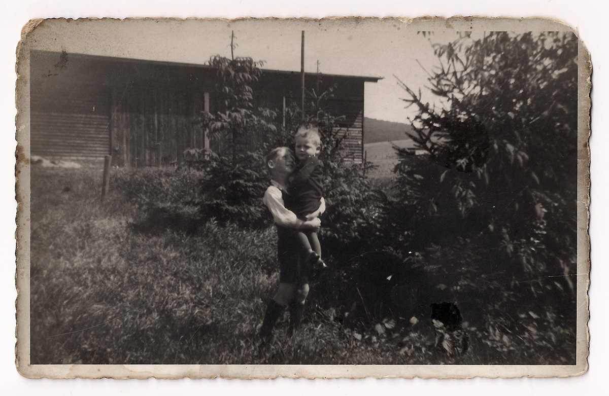 Es ist ein schwarz-weißes Foto. Zwei Jungen stehen in einem Garten. Der Ältere trägt den Jüngeren. Der Ältere blickt freundlich zu seinem Bruder. Dieser guckt in die Kamera.