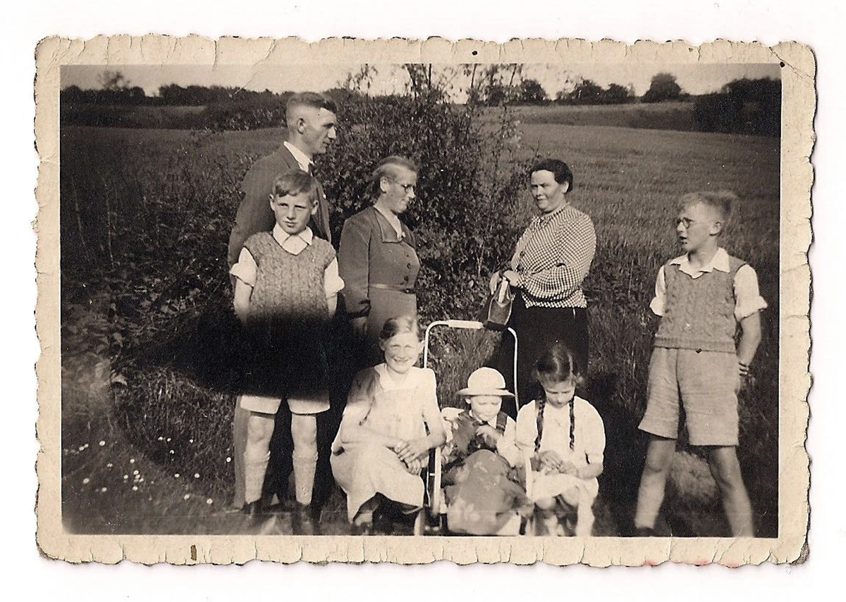 Es ist ein schwarz-weißes Foto. Die Familie steht auf einem Feld mit Büschen im Hintergrund. In einer Karre mit hohem Griff sitzt Heinz Schäfer mit einem runden Hut. Neben ihm sitzen zwei Mädchen in hellen Kleidern. Daneben stehen zwei Jungen in kurzen Hosen mit Pollunder. Hinter dem Wagen stehen ein großer mittelalter Mann, eine ältere Frau in Blusenkleid und eine jüngere Frau mit dunklem Rock und gemusterter Jacke.