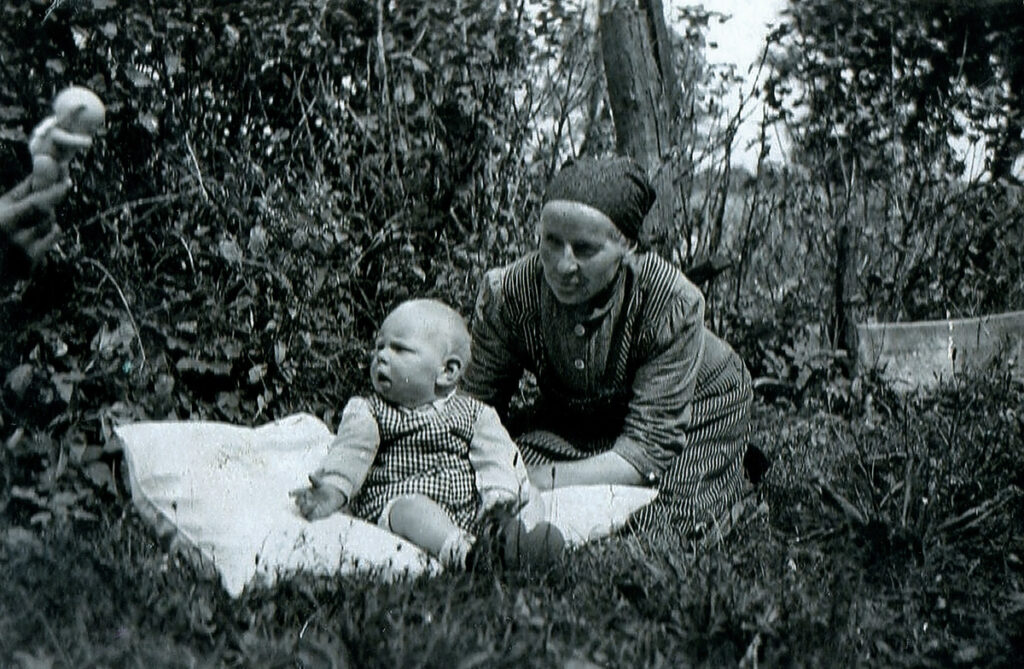 Es ist ein schwarz-weißes Foto. Fritz sitzt als Baby auf einer hellen Decke auf einer Wiese. Er wird im Sitzen gehalten. Seine Großmutter hält ihn. Er guckt erstaunt zum linken Bildrand. Dort ist eine Hand im Bild. Die Hand hält einen Teddybären Richtung Fritz. Die Großmutter trägt ein Kopftuch, ein Arbeitshemd und eine Schürze. Fritz trägt einen hellen Pulli und einen Strampelanzug.