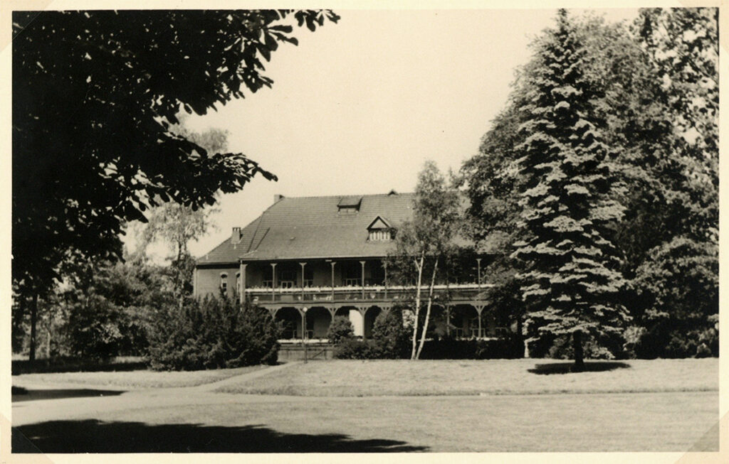 Es ist ein schwarz-weißes Foto von Haus 25. Es ist ein zweistöckiges Haus hinter hohen Bäumen. Das Haus hat auf der gesamten Längsseite des Gebäudes hölzerne Balkone mit Überdachung.
