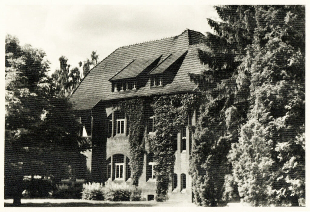 Es ist ein schwarz-weißes Foto von Haus 23. Das Haus ist zweistöckig. Es ist umgeben von hohen Bäumen. Am Gebäude wächst eine Rankpflanze bis ans Dach heran.