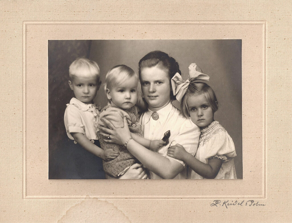 Es ist ein schwarz-weißes Foto. Elly Endewardt trägt Jürgen auf dem Arm. Dieter und Ute halten die beiden. Elly trägt eine helle hochgeschlossene Bluse mit einer Brosche am Hals. Ihre Haare sind sorgfältig hochsteckt. Sie blickt freundlich in die Kamera. Die Kinder tragen helle Kleidung und blicken ebenfalls zur Kamera. Ute trägt eine große Schleife im Haar.