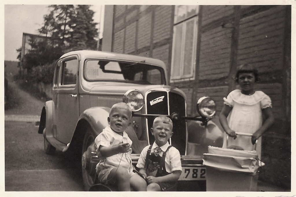 Es ist ein Schwarz-Weiß-Foto. Die Kinder stehen vor einem Opel Rekord. Heinrich und Helmut tragen helle Hemden, Irmgard ein helles Kleid. Sie schiebt außerdem einen Puppenwagen vor sich. Alle drei Kinder gucken fröhlich.