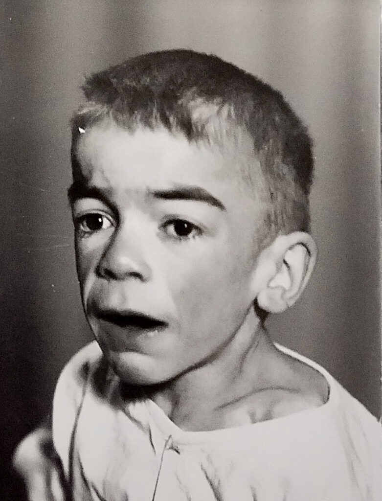 Es ist ein schwarz-weißes Fotoporträt von dem Jungen. Er trägt ein weißes Hemdchen. Hinter ihm ist ein Bühnenvorhang zu erkennen.
