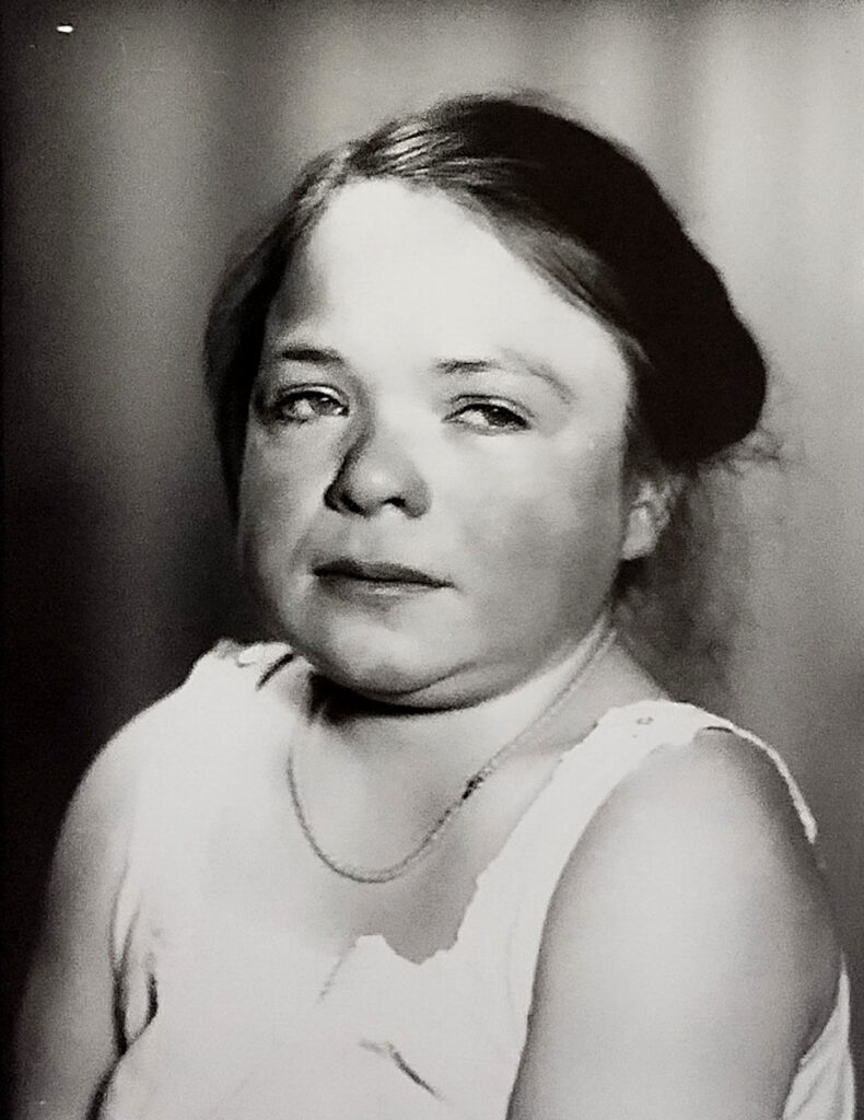 Es ist ein schwarz-weißes Porträtfoto. Zu sehen ist ein Mädchen. Sie trägt ein helles Hemd und eine Halskette. Ihre Haare sind zusammengebunden. Sie hat geweint. Sie schaut nicht zur Kamera und ihr Blick ist traurig.