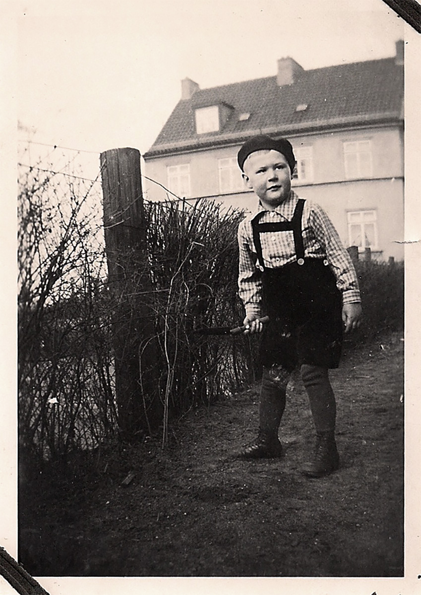 Es ist ein schwarz-weißes Foto. Hans Buhlrich steht in einem Garten vor einem Zaun. Er ist etwa vier Jahre alt. Er trägt eine dunkle kurze Latzhose mit kariertem Hemd und eine Mütze. Er hält eine Schaufel in der rechten Hand und schaut nach links aus dem Bild heraus.