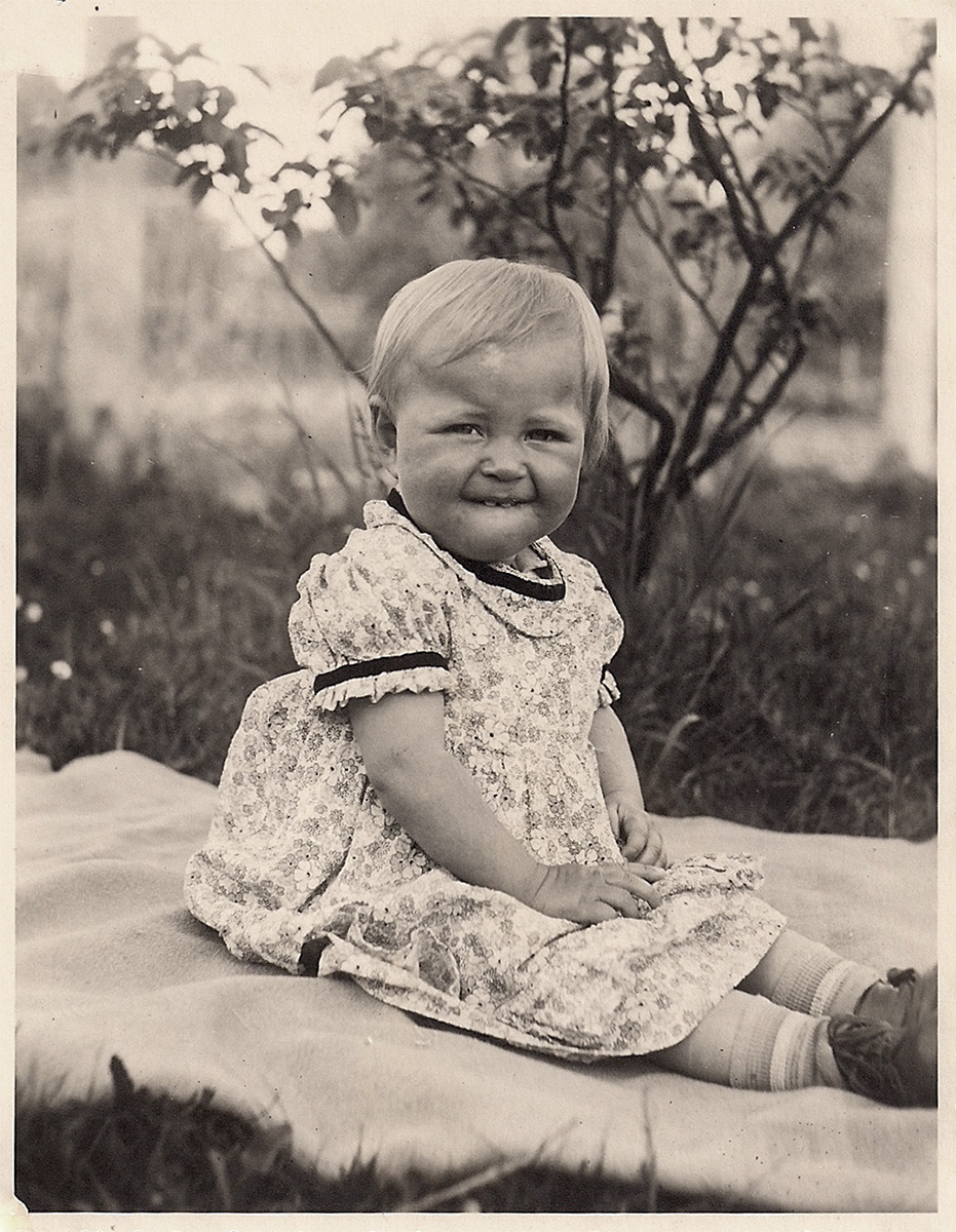 Es ist ein schwarz-weißes Foto. Erika ist etwa ein Jahr alt. Sie sitzt auf einer Decke in einem Garten. Sie trägt ein kurzes geblümtes Kleid. Sie beißt auf ihre Unterlippe und guckt interessiert zur Kamera.