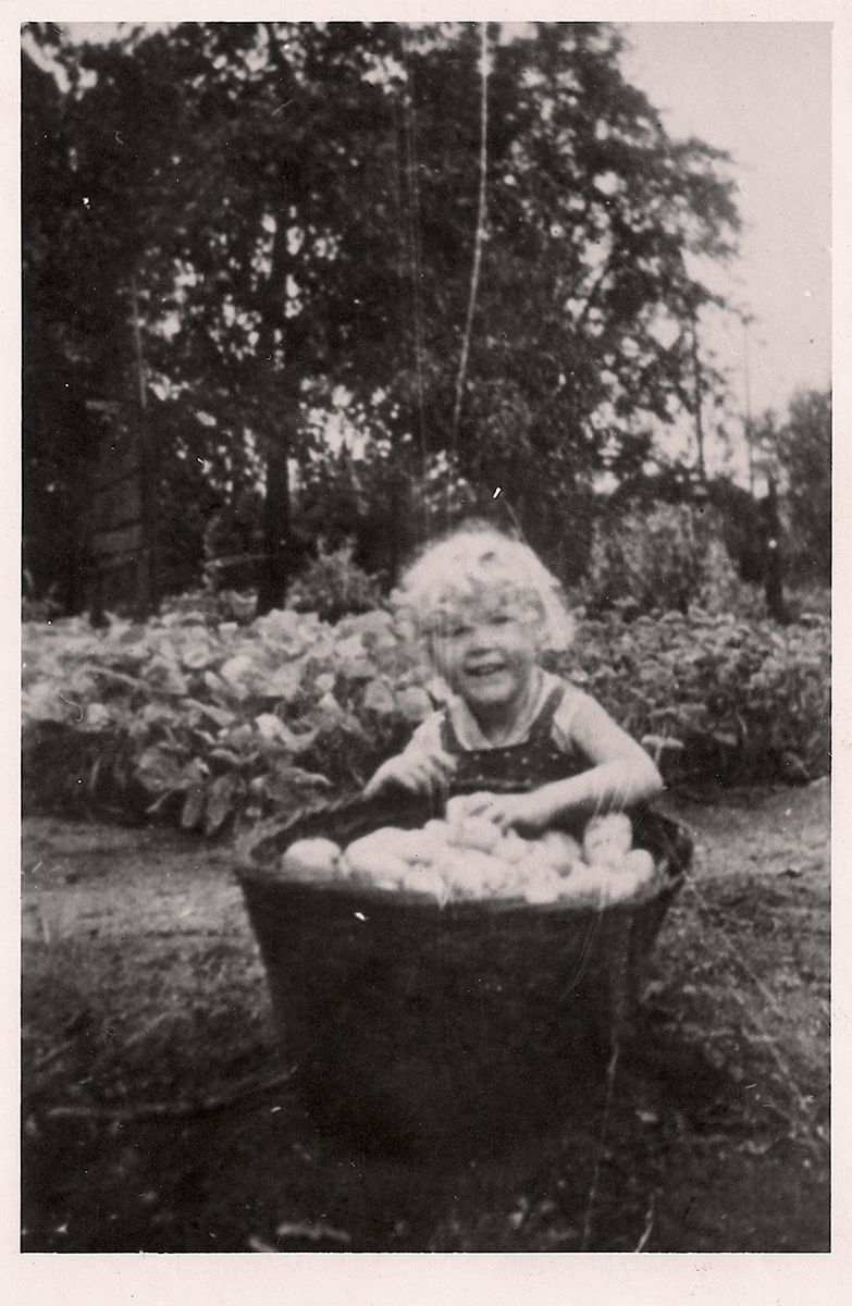 Es ist ein schwarz-weißes Foto. Margret sitzt in einem Garten hinter einem großen Korb mit Äpfeln. Sie hat helle Locken und blickt lachend zur Kamera. Sie trägt ein ärmelloses Hemdchen und ist etwa drei Jahre alt.