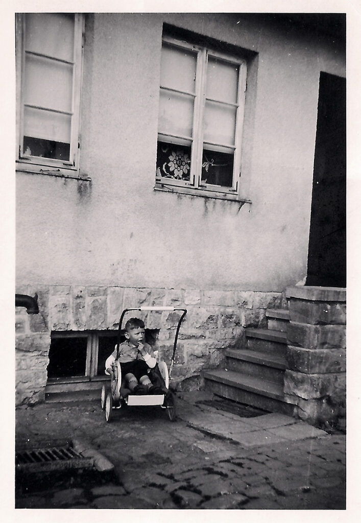 Es ist ein schwarz-weißes Foto. Zu sehen ist ein Kinderwagen, der vor einem verputzten Haus steht. Im Kinderwagen sitzt ein Kind. Es trägt kurze Hosen und ein Hemd. Es scheint etwas am Rand zu betrachten und guckt zur Seite.