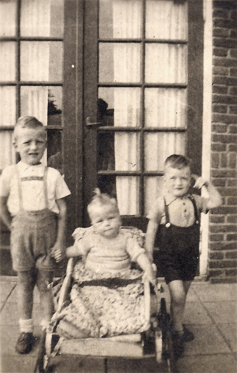 Es ist ein schwarz-weißes Foto. Es ist leicht unscharf. Drei Kinder stehen vor einer Sprossentür. In der Mitte sitzt ein Kind in einem Wagen. Zu jeder Seite steht ein weiterer Junge. Die Jungen tragen kurze Latzhosen. Das Kind im Wagen trägt ein helles Oberteil und einen Zopf oben auf dem Kopf.