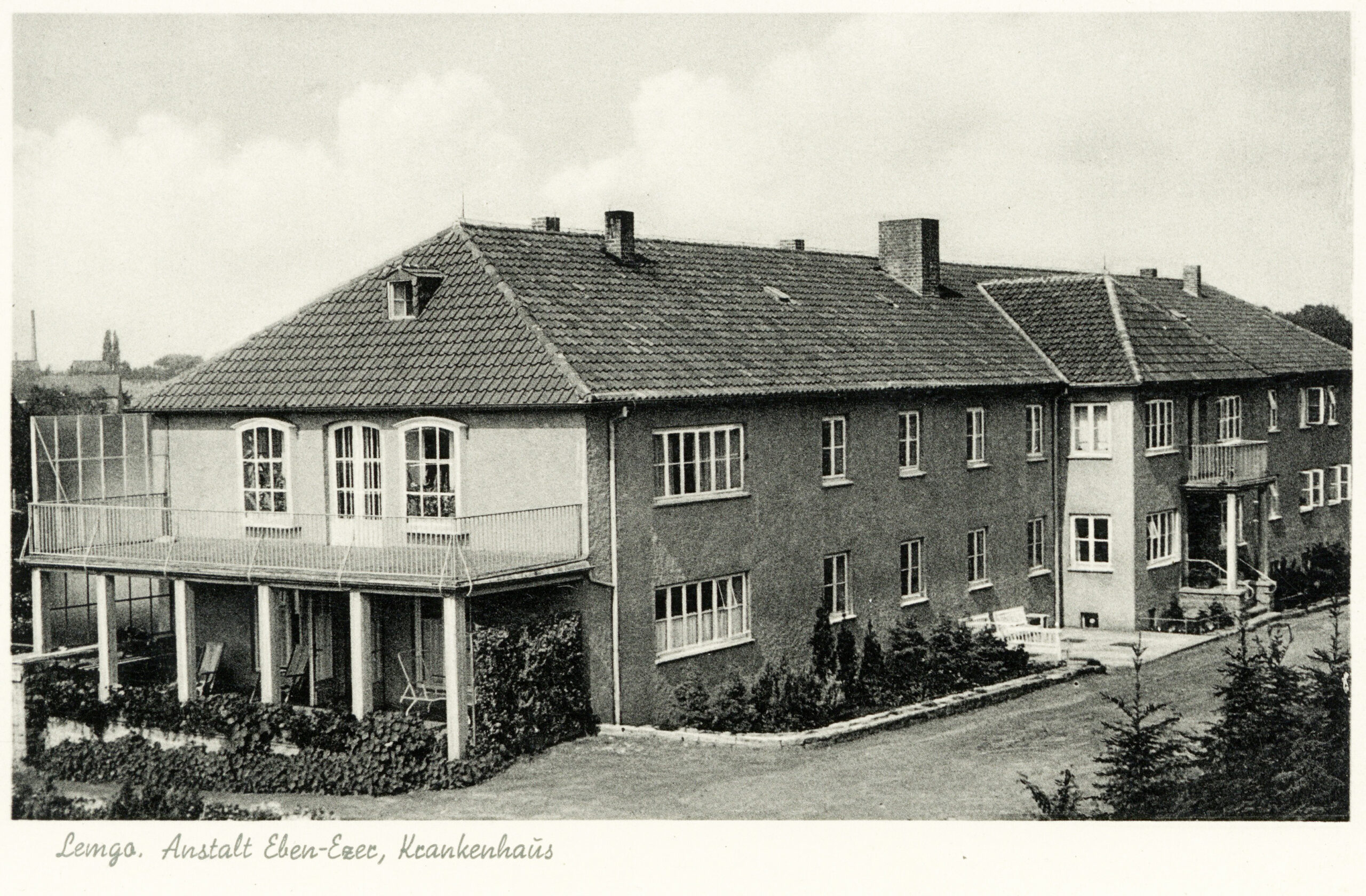Es ist eine schwarz-weiße Postkarte des Krankenhausgebäudes der Anstalt Eben-Ezer in Lemgo. Das breite Gebäude mit Spitzdach ist zweigeschossig und hat zur einen Giebelseite einen breiten Balkon, der mit sechs Pfeilern abgestützt wird und unter dem sich eine Terrasse befindet. Auch über dem mittigen Haupteingang zum Gebäude ist ein Erker mit einem Balkon, von Säulen abgestützt.