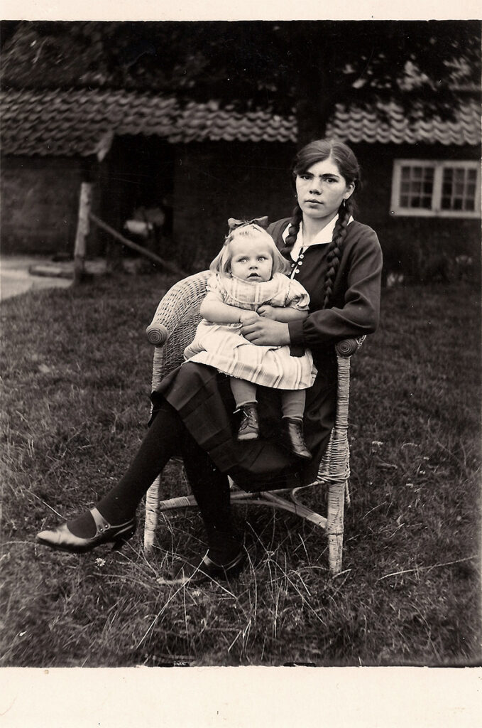 Das ist ein Schwarz-Weiß-Foto. Hilda sitzt in einem Korbstuhl in einem Garten. Sie trägt ein dunkles Kleid und lange geflochtene Zöpfe. Nach rechts angelehnt blickt sie in die Kamera. Auf ihrem Schoß sitzt seitlich Frieda. Frieda trägt ein helles kurzärmeliges Kleid und hält eine Hand von Hilda. Frieda guckt nach links aus dem Bild heraus.