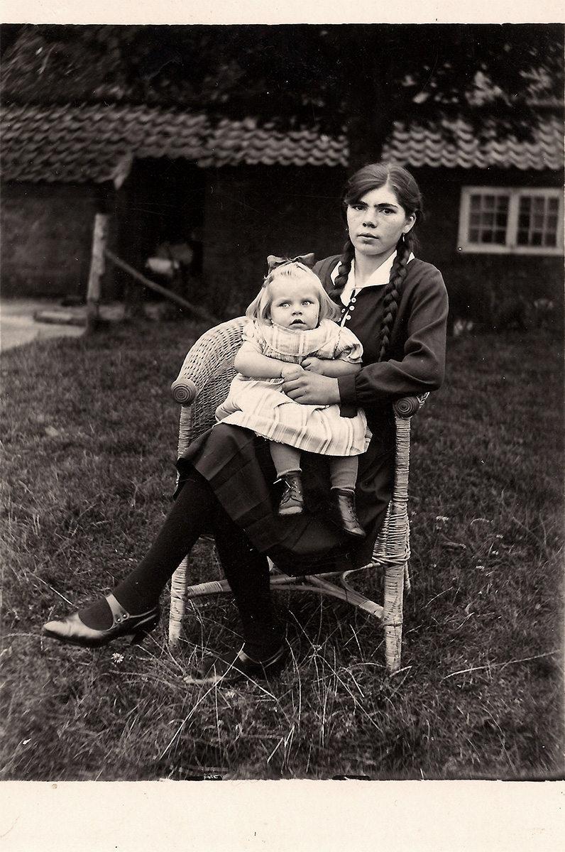 Das ist ein Schwarz-Weiß-Foto. Hilda sitzt in einem Korbstuhl in einem Garten. Sie trägt ein dunkles Kleid und lange geflochtene Zöpfe. Nach rechts angelehnt blickt sie in die Kamera. Auf ihrem Schoß sitzt seitlich Frieda. Frieda trägt ein helles kurzärmeliges Kleid und hält eine Hand von Hilda. Frieda guckt nach links aus dem Bild heraus.
