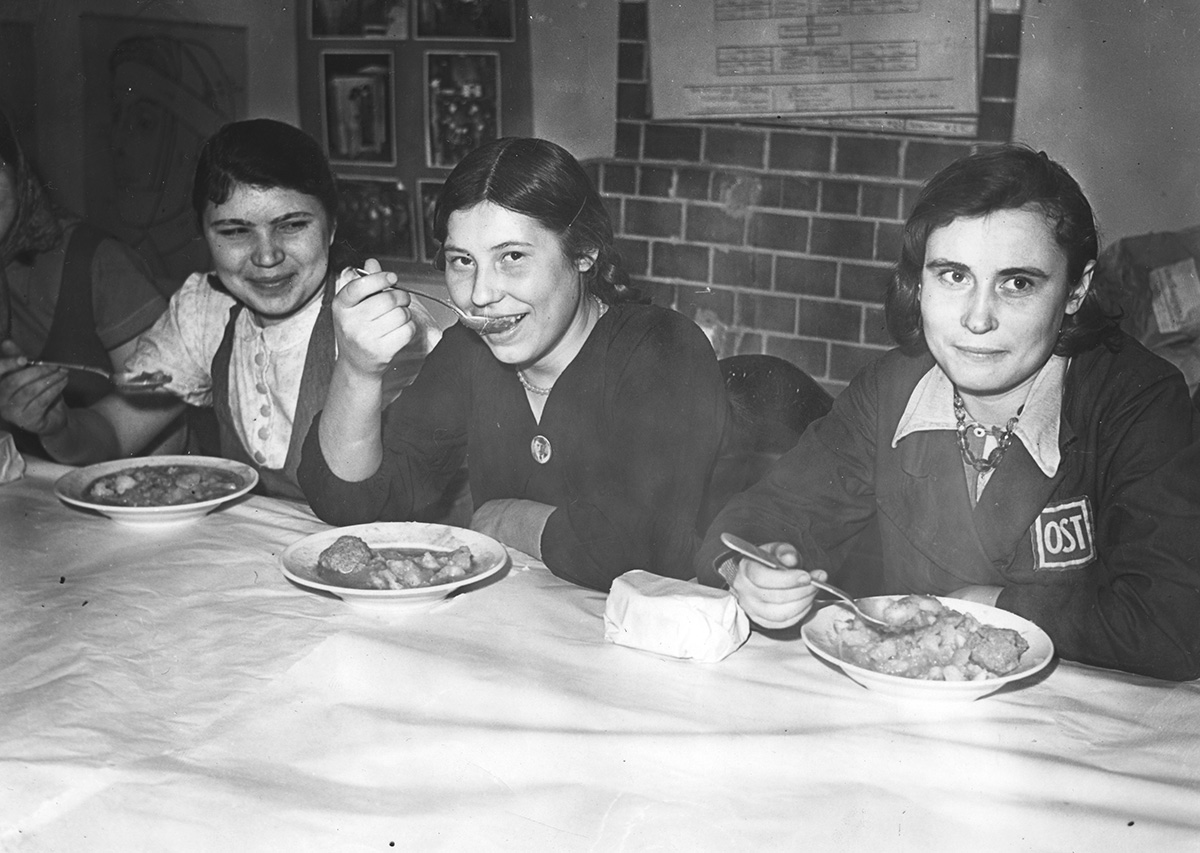 Die drei Frauen blicken fröhlich. Die Teller sind reichlich gefüllt. Sie sind sorgfältig frisiert und tragen unterschiedliche Kleidung. Auf der Kleidung der rechten Frau ist ein Aufnäher mit dem Begriff »OST« zu sehen.