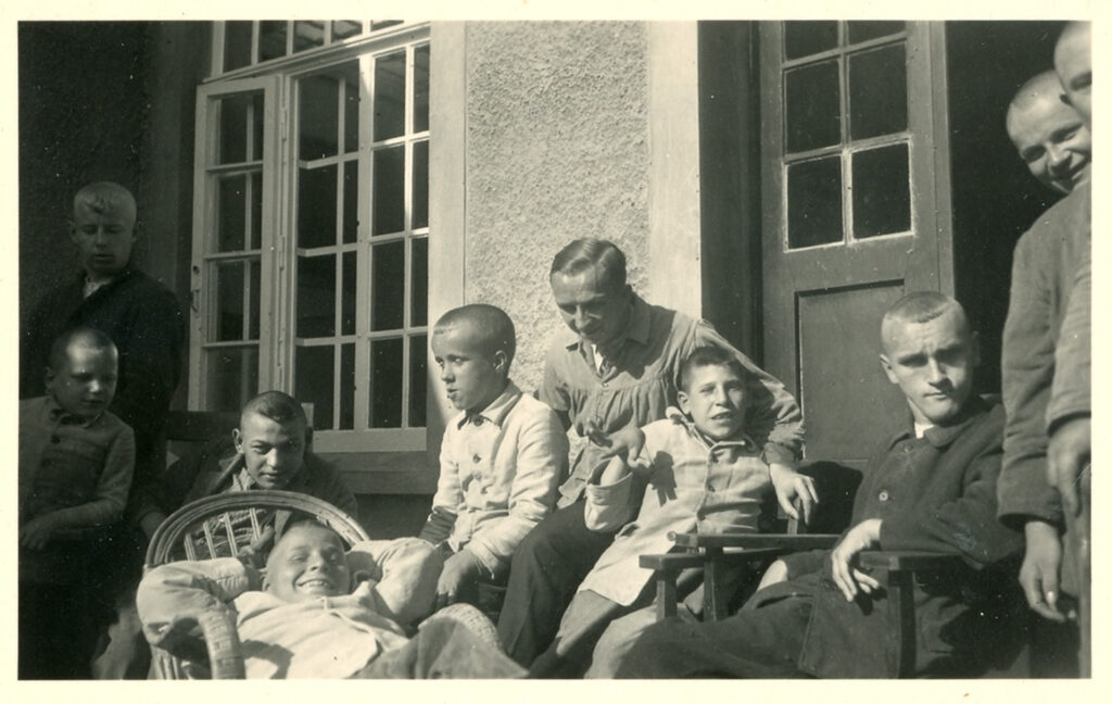 Es ist ein schwarz-weißes Foto mit zehn Kindern. Die Kinder sind unterschiedlich alt, ungefähr eineinhalb bis dreizehn Jahre. Sie sitzen auf unterschiedlichen Stühlen. Es ist ein spontanes Bild, nicht arrangiert. Bei den Kindern sitzen drei Frauen in Arbeitskleidung der Pflegerinnen.