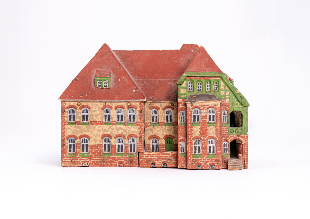 Die Miniatur ist aus Holz. Es ist ein Nachbau des Hauses 24. Es ist ein zweigeschossiges Gebäude. Die Ziegel sind rot, grau und gelb. Über den Fenstern gibt Muster im Mauerwerk. Es gibt einen zweigeschossigen Erker mit Balkon und ein Fachwerkgiebel. Der Erker und die Balken im Giebel sind in einem hellen Grünton. Das Dach ist ein rotes Spitzdach. Die Bauart ist von Neunzehnhundert.