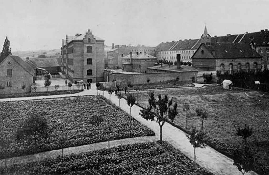 Es ist ein Schwarz-Weiß-Bild. Hinter gerade angelegten Beeten mit einer kleinen Allee ist das Zellenhaus der Anstalt. Es hat drei Stockwerke und drei kleine Stufengiebel über den Eingängen. Es liegt eingebunden zwischen ähnlichen Gebäuden in dem Anstaltskomplex.