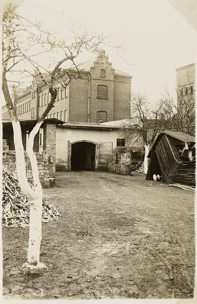 Es ist ein Schwarz-Weiß-Bild eingeklebt in eine Fotoalbum. Der Schuppen ist einstückig und hell verputzt. Er hat ein zweiflügeliges Tor. Die Tore sind weit geöffnet. Vor dem Schuppen ist eine freie Fläche, auf der ein Stapel Holz und Steine erkennbar sind. Im Hintergrund ist das Zellenhaus zu erkennen.
